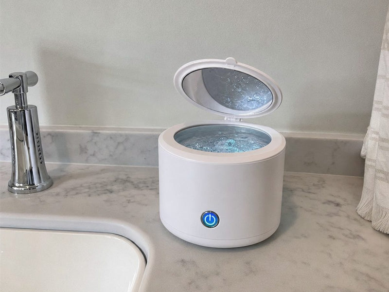 The Rinsa pod on a light stone bathroom counter beside a chrome tap, lid open and the water inside full of bubbles.