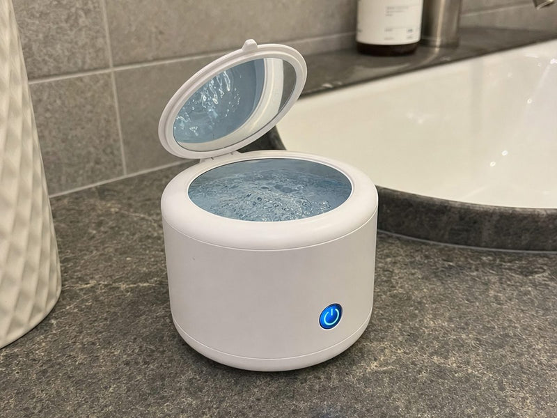 The Rinsa pod on a grey stone bathroom counter next to a basin, lid open and the water inside moving.